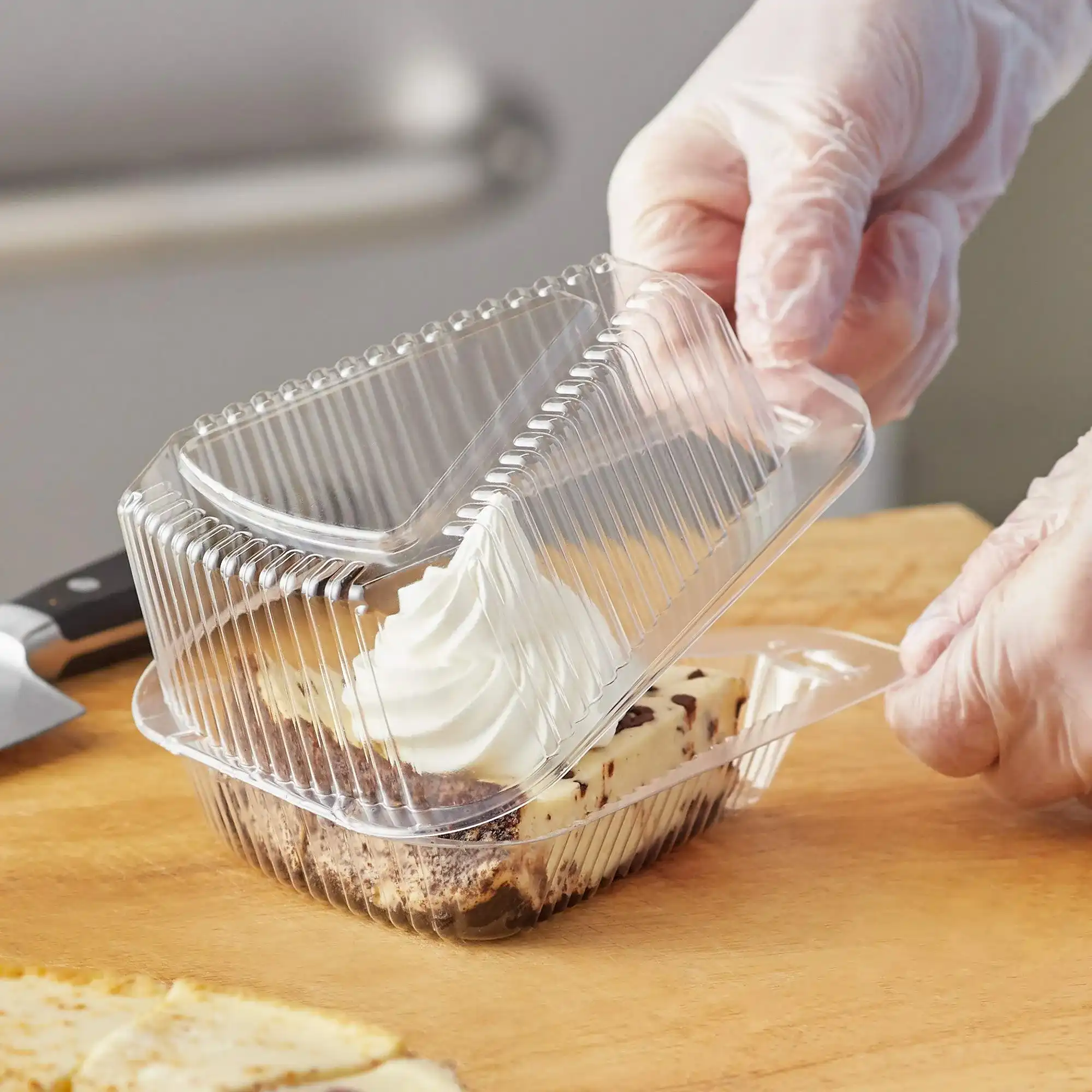 A person wearing gloves opening a clear plastic hinged container with a slice of cake inside, topped with whipped cream.