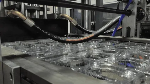Primer plano de una termoformadora que forma envases de plástico transparente, con mangueras hidráulicas visibles y moldes metálicos en un entorno industrial.