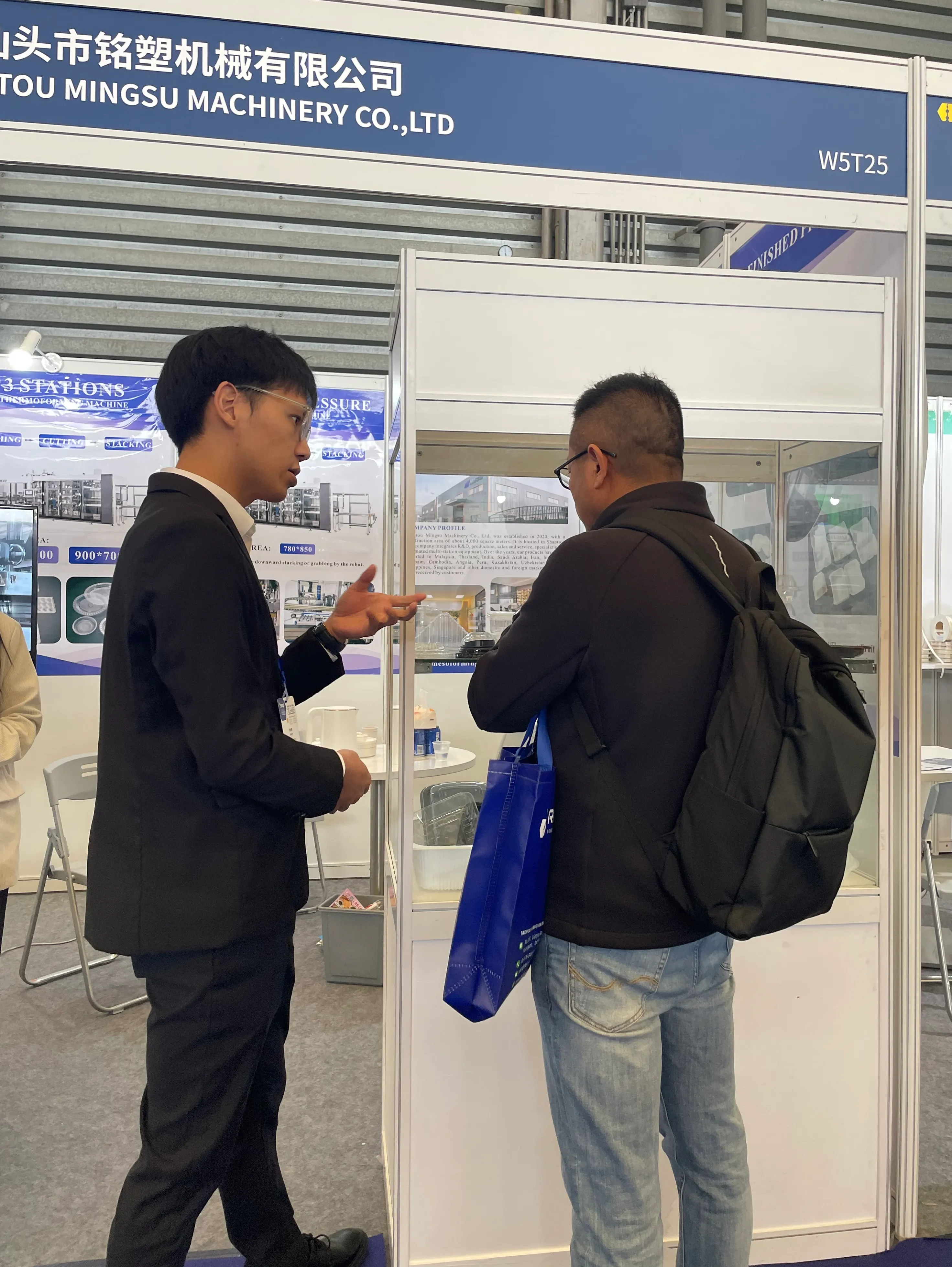 MingSu Machinery Co., Ltd. representative discussing thermoforming machinery with a visitor at an exhibition booth.