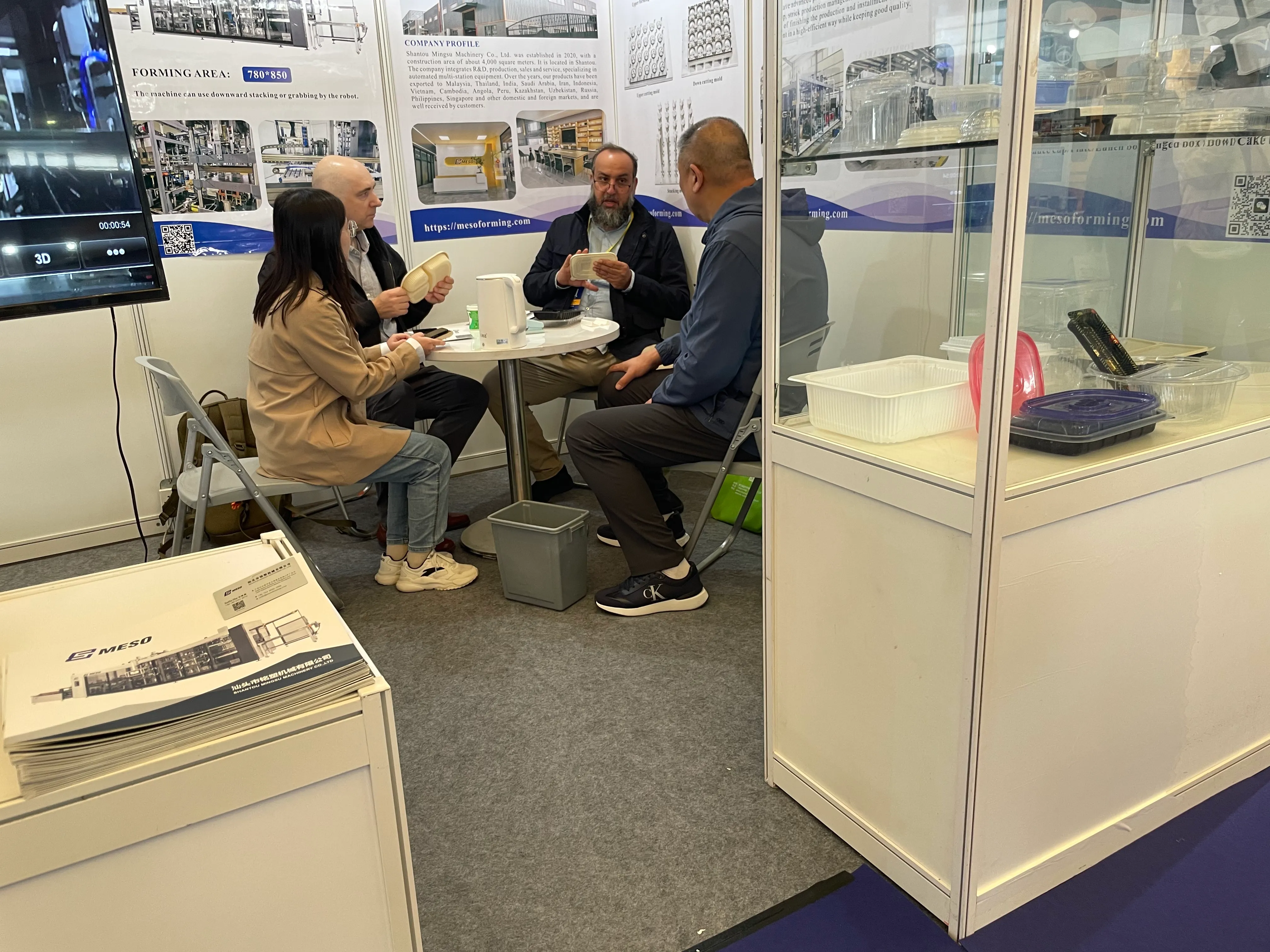 Four individuals seated at a table in a trade show booth discussing plastic packaging products.