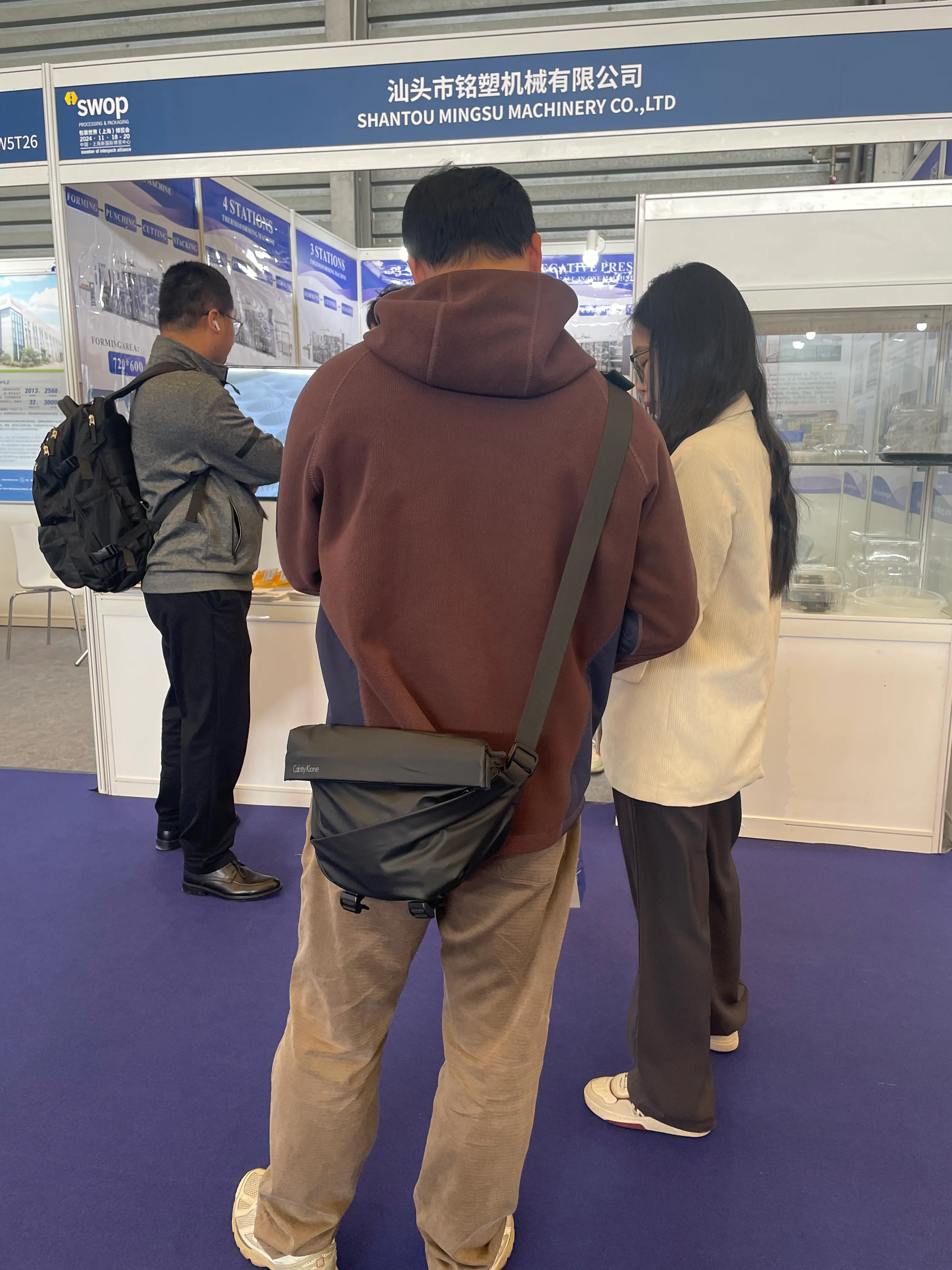 Three individuals standing in front of a trade show booth for Shantou Mingsu Machinery Co., Ltd.